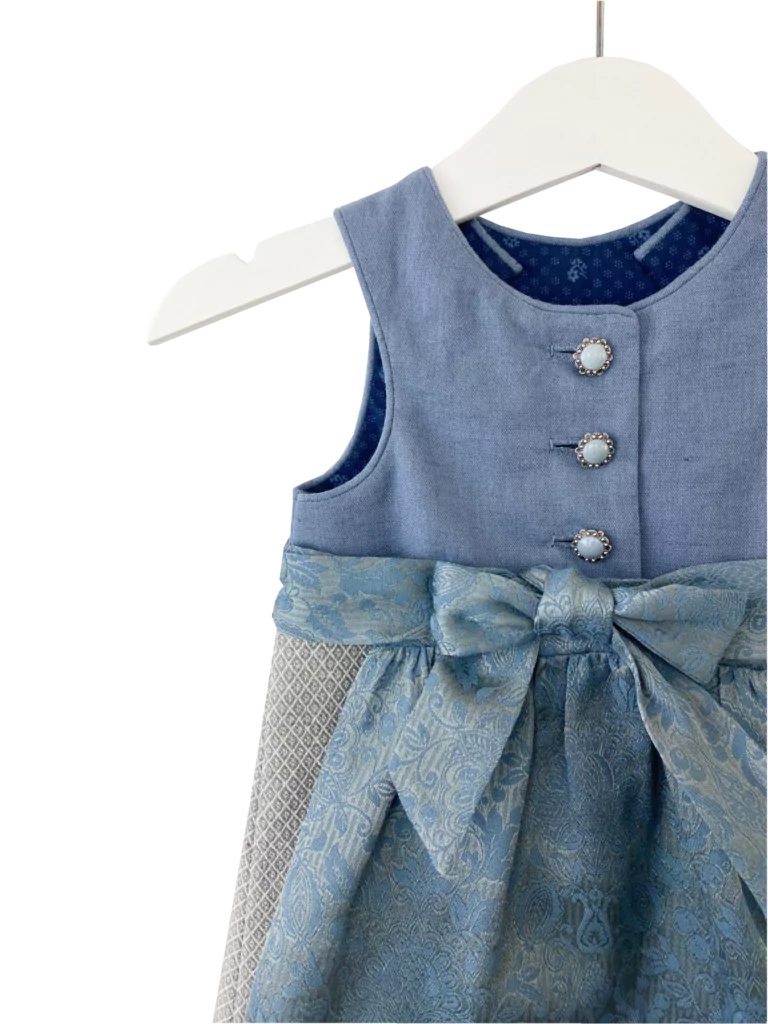 Babydirndl aus blauem Leinenoberteil mit passepoilliertem Ausschnitt und 3 Knöpfe. Gemusterter Leinenrock mit Falten. Schürze ist gemustert mit Bund und Bänder zum binden.