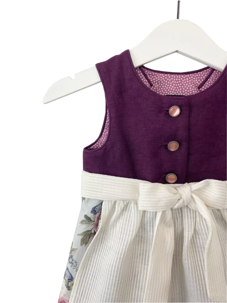 Babydirndl aus bordeaux-farbenen Leinenoberteil mit passepoilliertem Ausschnitt und 3 Knöpfe. Geblümter Rock mit Falten. Schürze ist einfärbig creme mit Bund und Bänder zum binden.