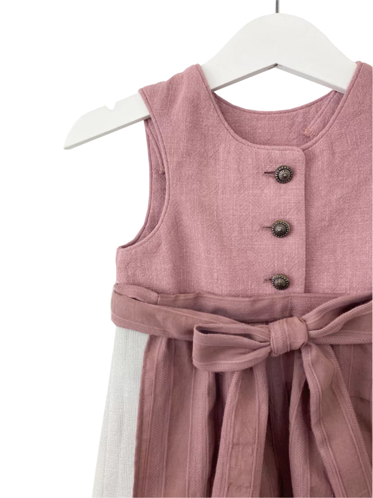 Babydirndl aus rosa-farbenen Leinenoberteil mit passepoilliertem Ausschnitt und 3 Knöpfe. Einfärbig beiger Rock mit Falten. Schürze ist einfärbig altrosa mit Bund und Bänder zum binden.