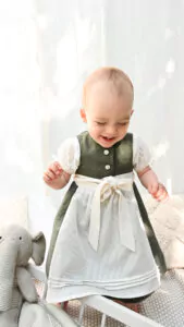 Babydirndl aus oliv-farbenen Leinenoberteil mit passepoilliertem Ausschnitt und 3 Knöpfe. Oliv-farbener Leinenrock gemustert mit Falten. Schürze ist creme-farben mit Bund und Bänder zum binden.